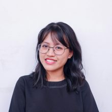 Portrait of a young woman with glasses and black hair smiling against a plain background.